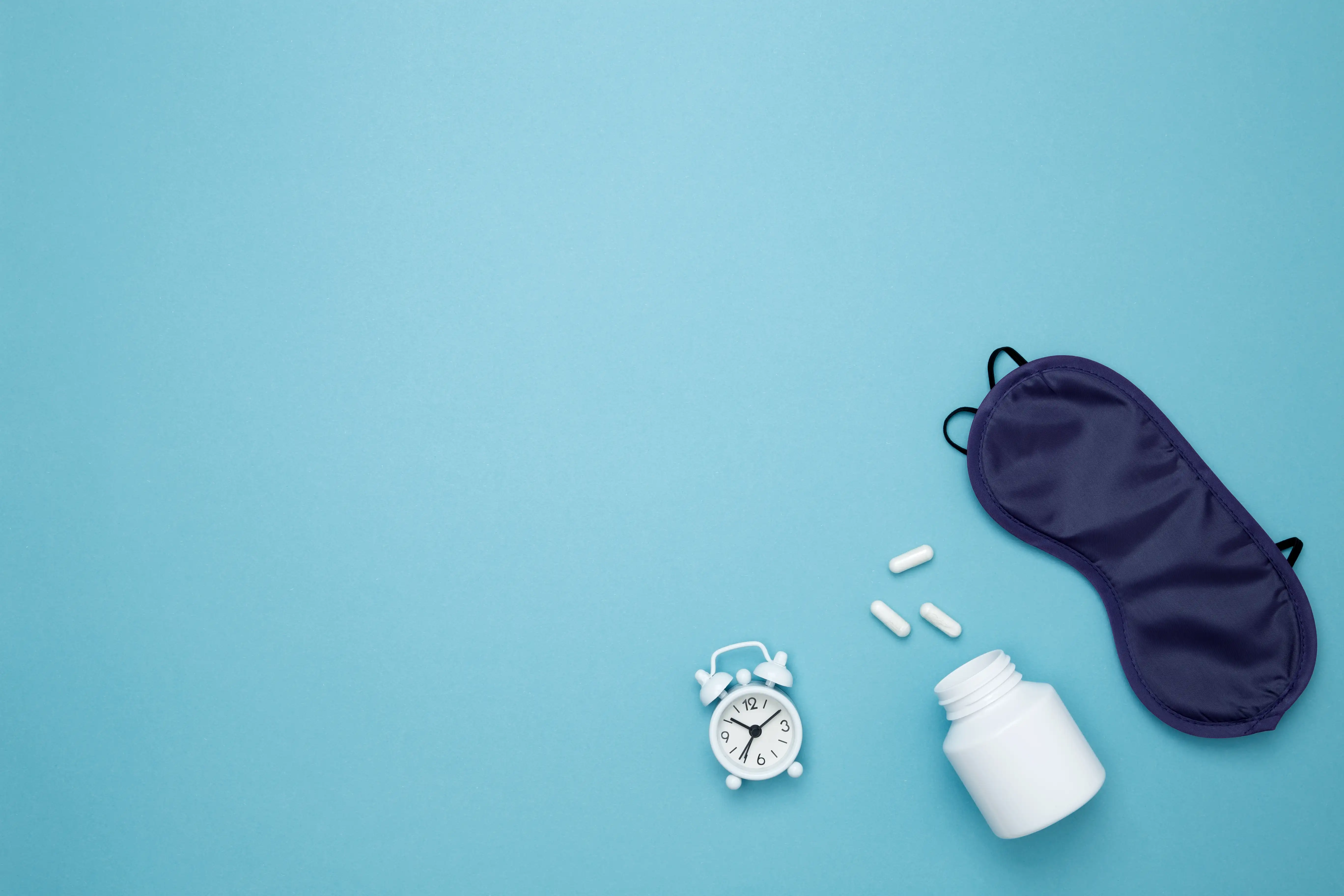 Sleep mask, capsules, and an alarm clock arranged on a tabletop