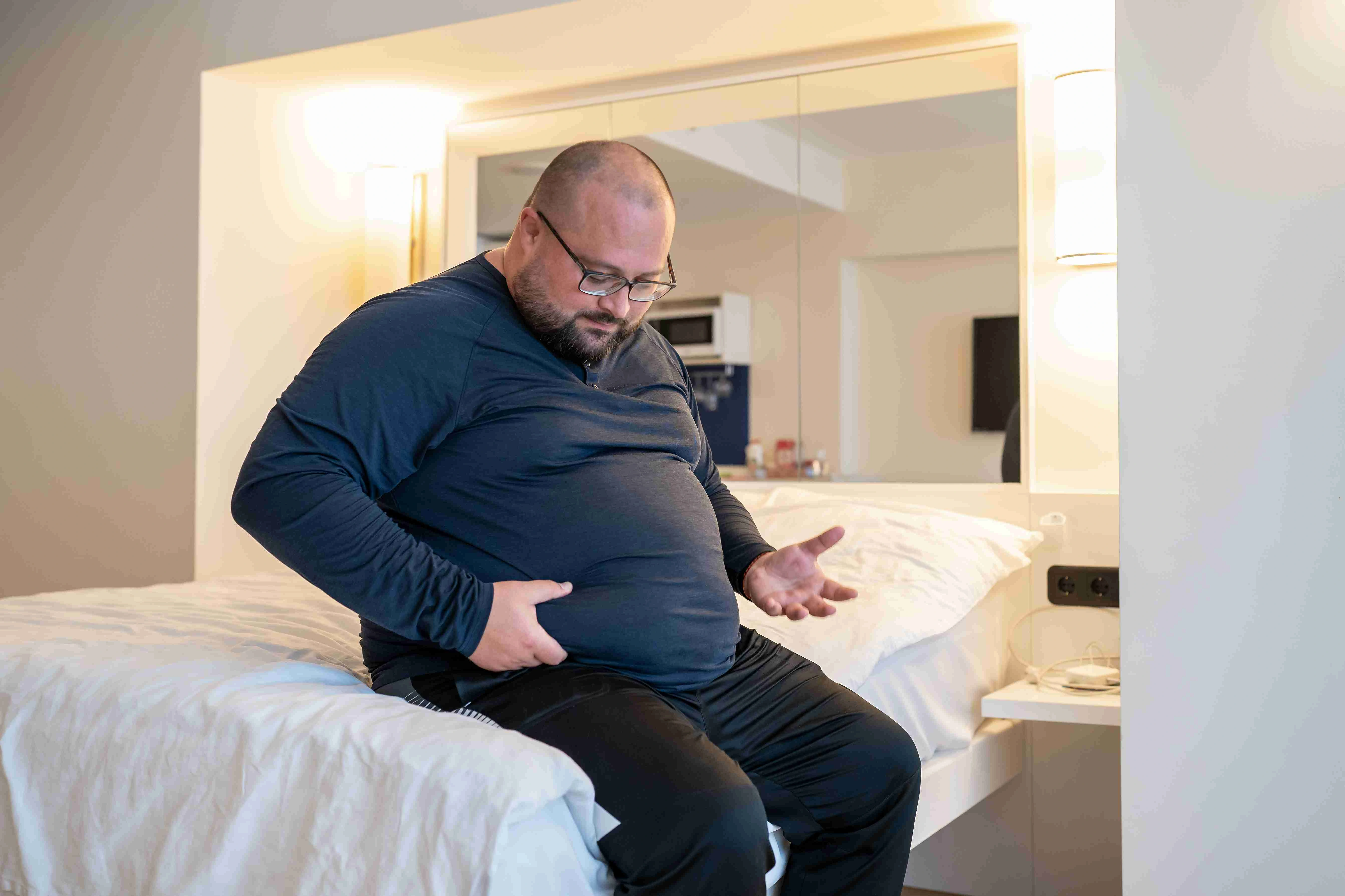 Huge man suffering from extra weight touching stomach sitting on bed in bedroom.