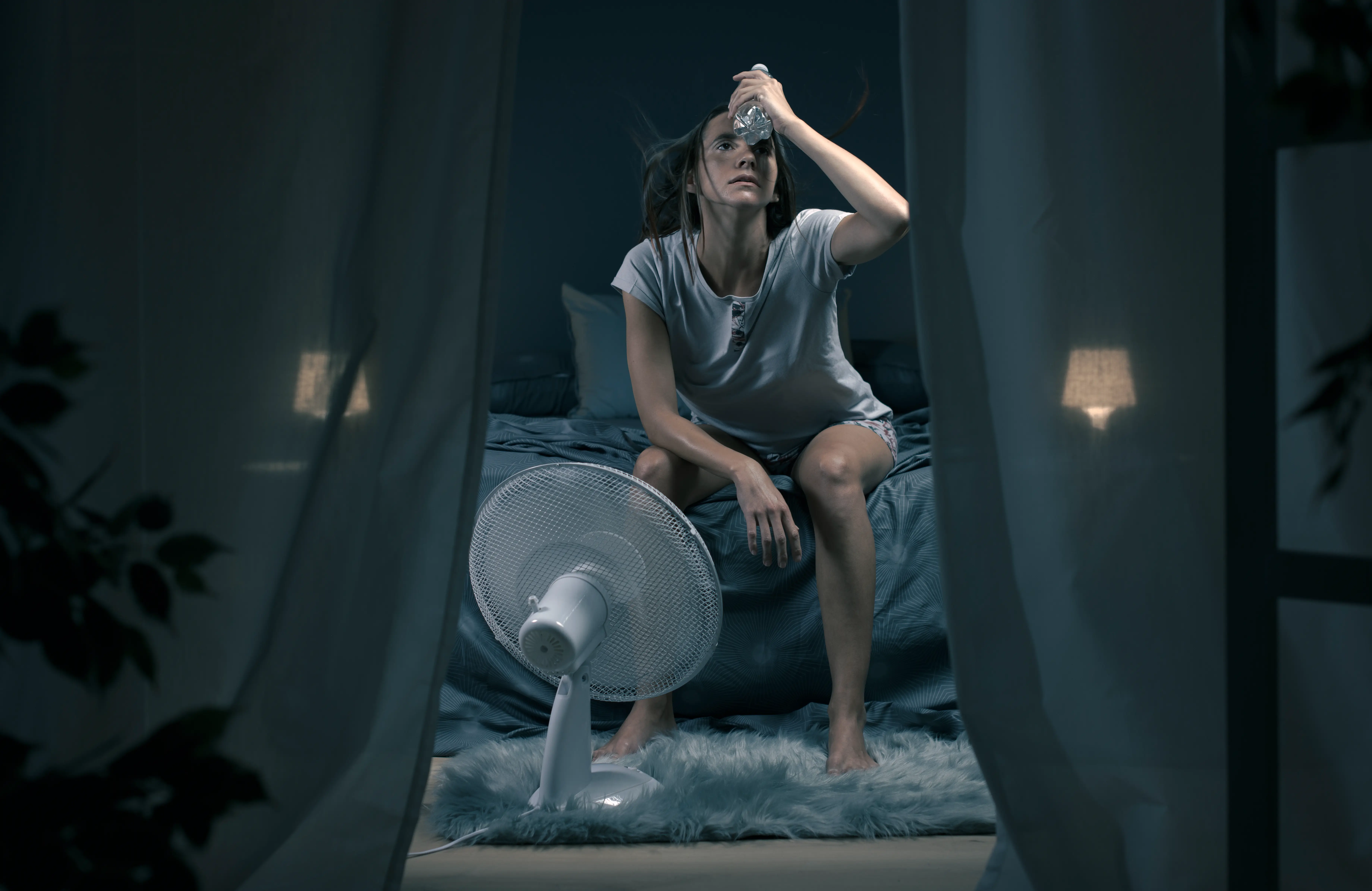 A woman holding a water bottle and sitting in front of a cooling fan in the bedroom