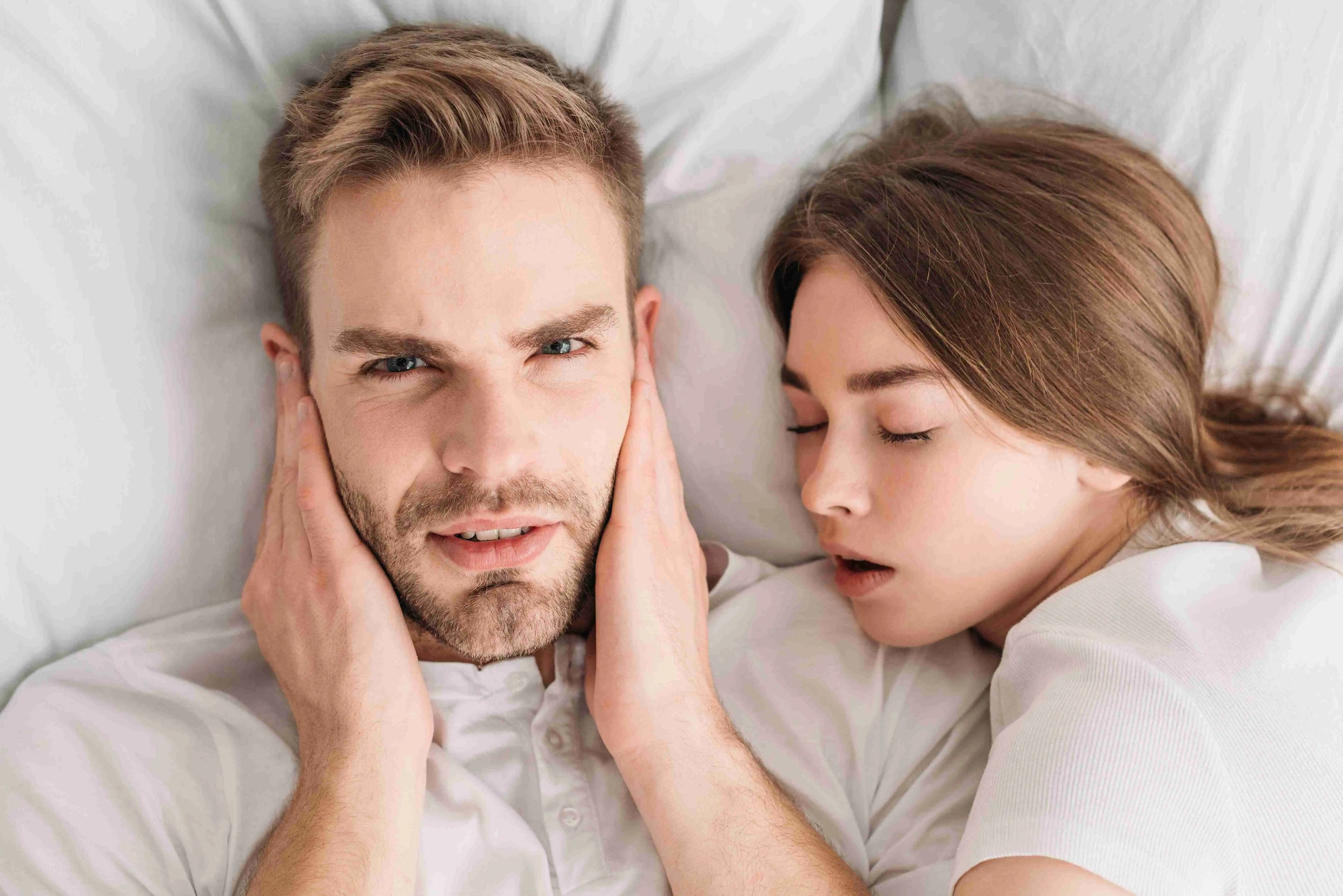 displeased man plugging ears with hands and looking at camera while lying in bed near snoring wife