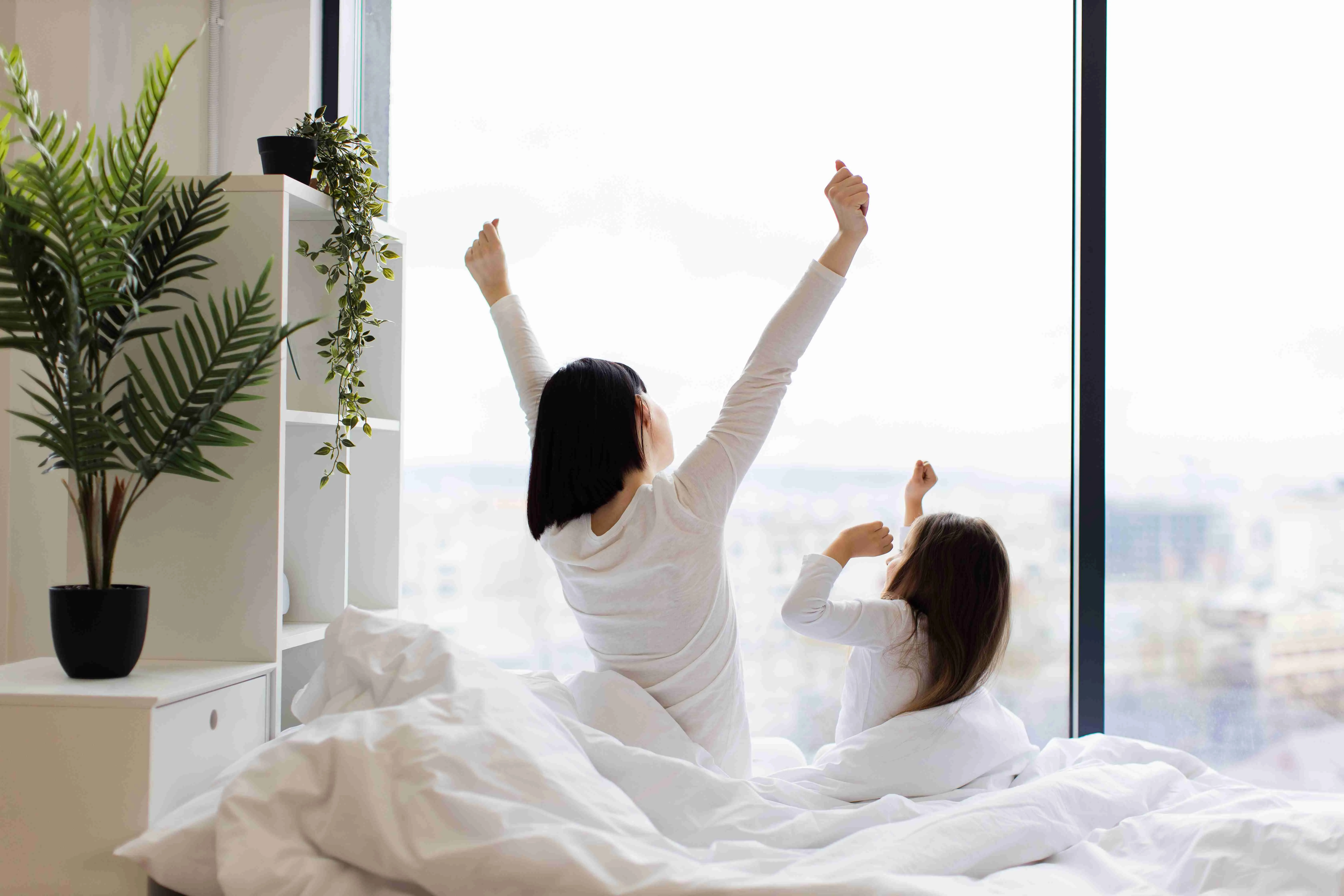 Mother and daughter stretching in bed in bright morning light by large window