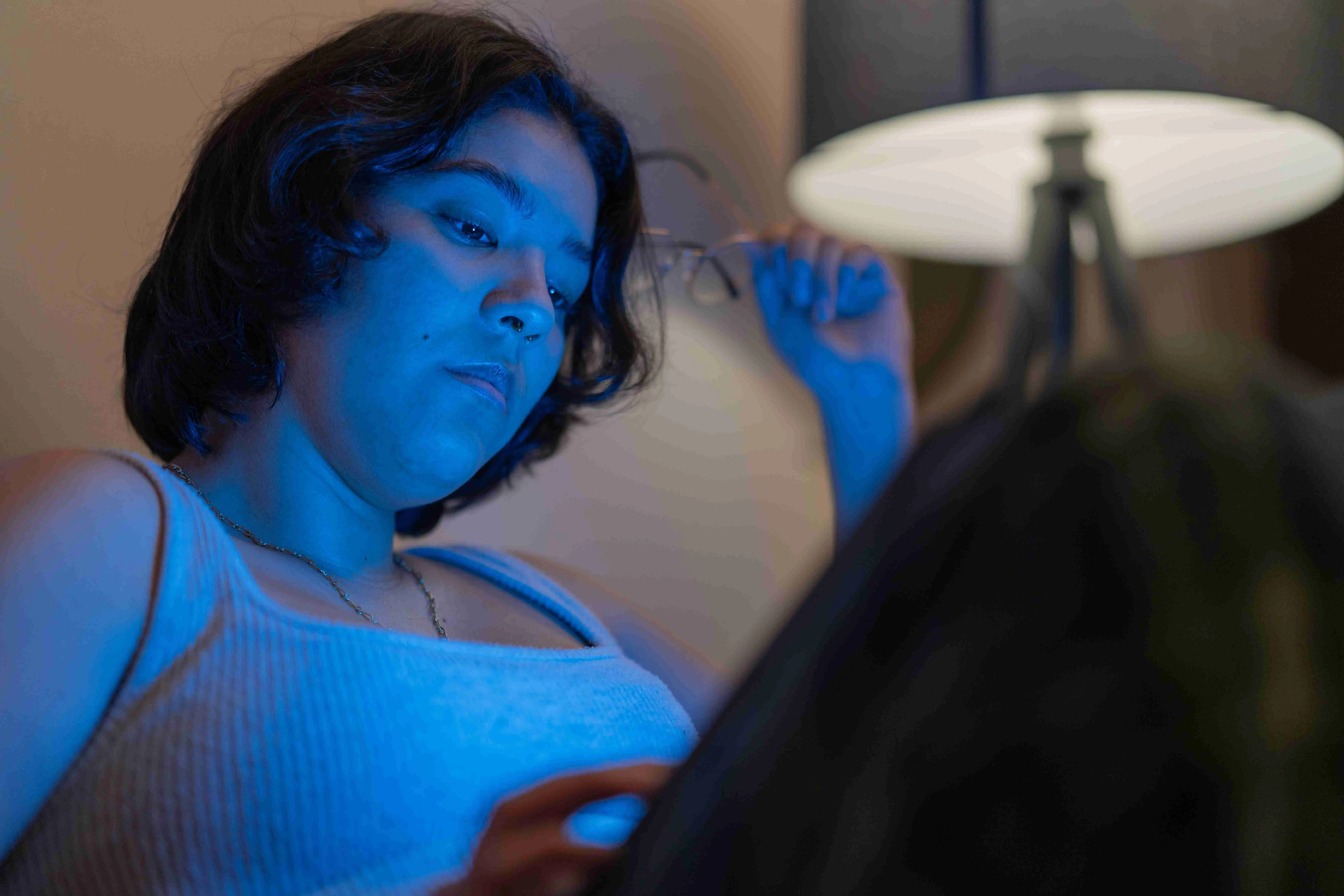 Young woman removes her glasses while reading on a tablet at night, illuminated by the blue light, suffering from insomnia and fatigue