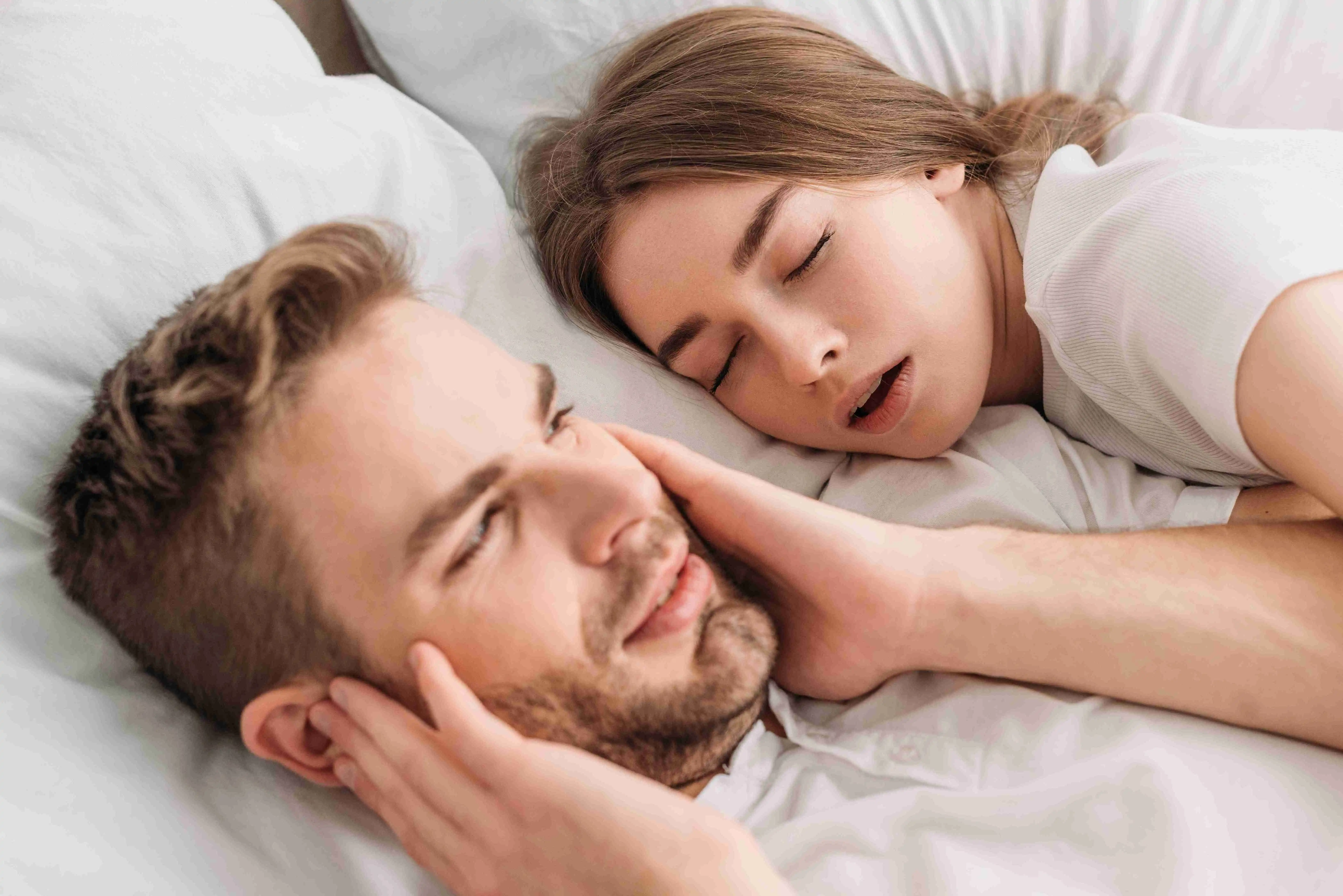 exhausted man plugging ears with hands while lying in bed near snoring wife