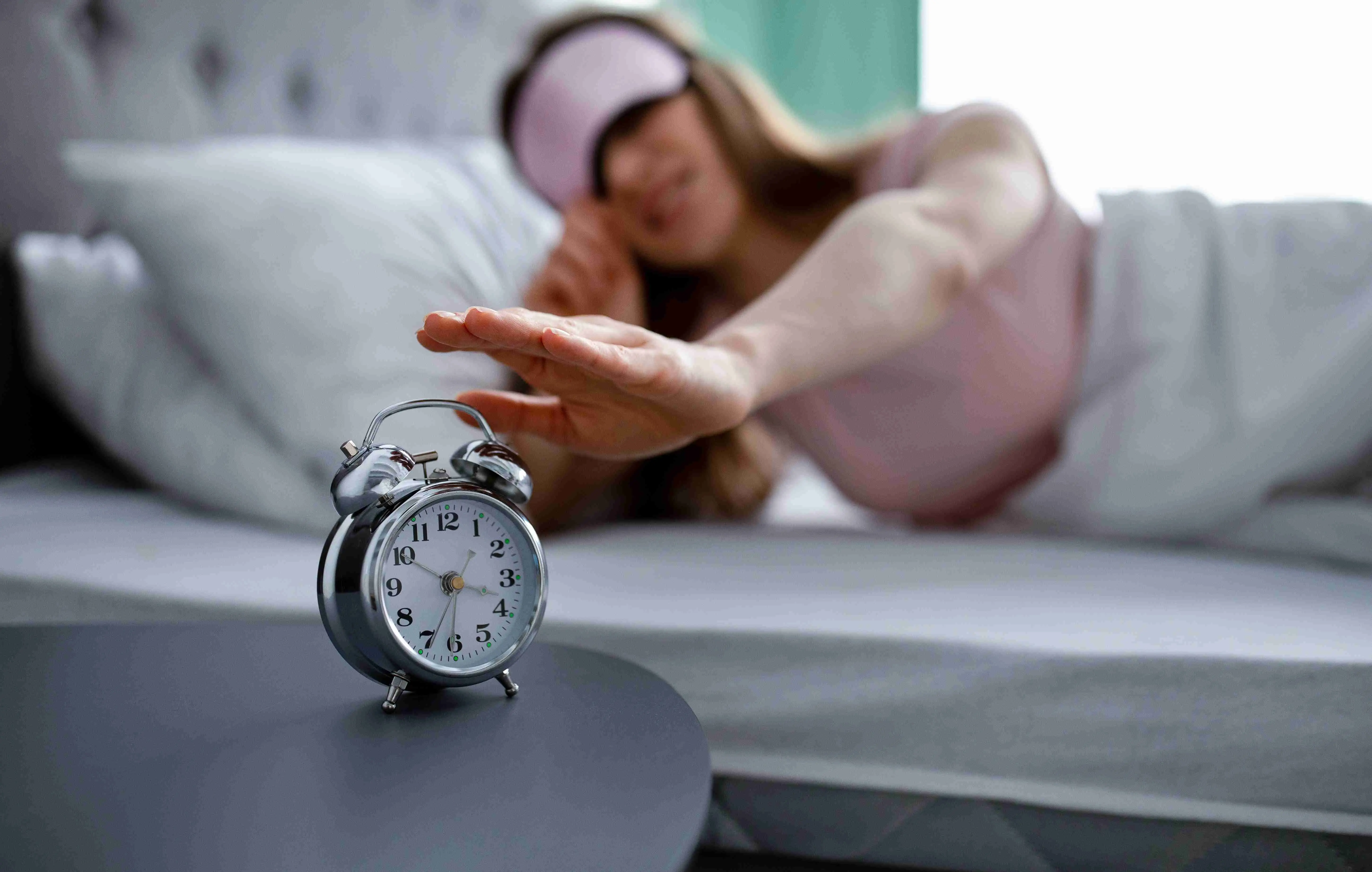 Young woman in sleep mask turning off alarm clock in morning