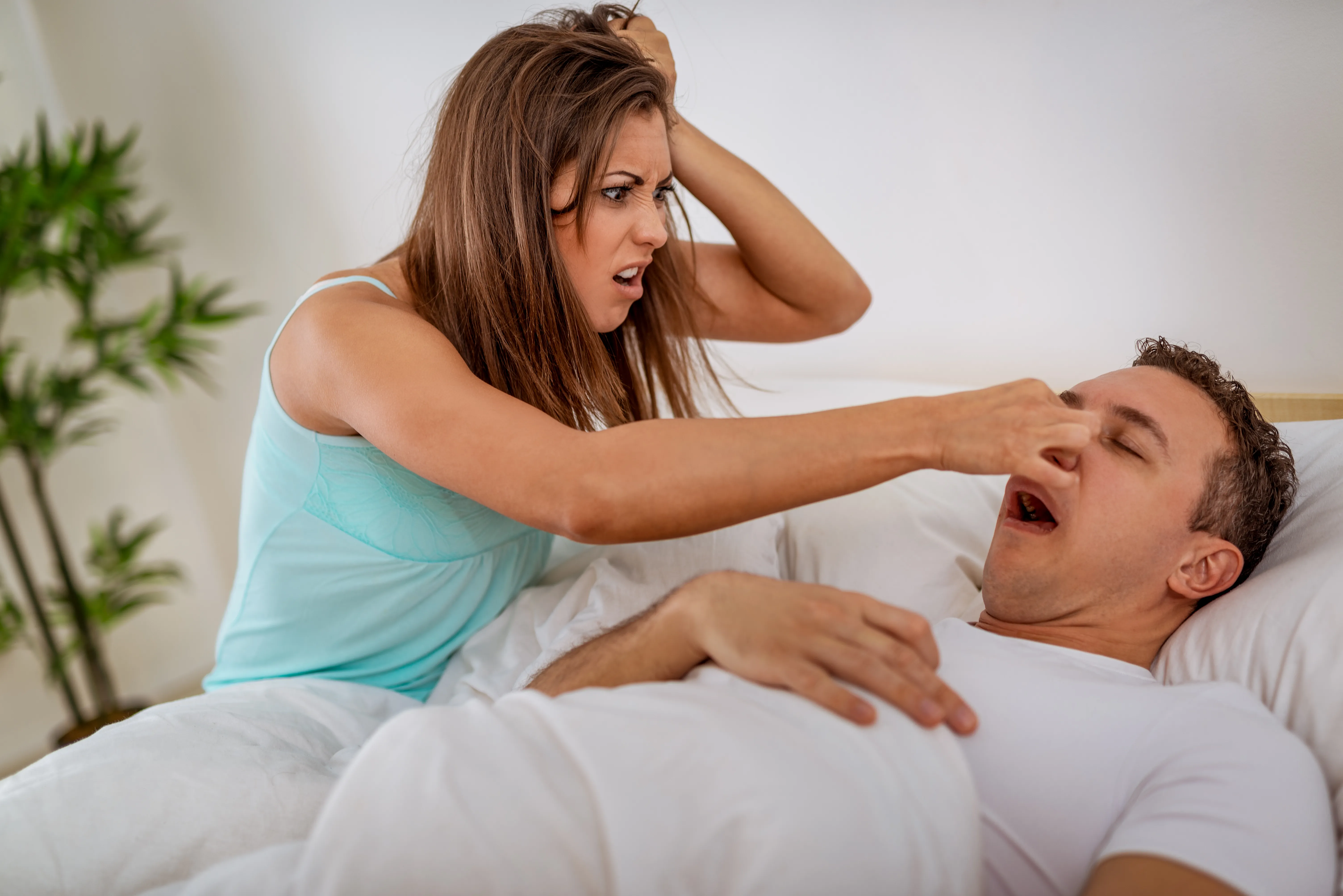 man sleeps and snores with mouth open, while a tired woman, irritated by snoring, sits on bed and pinches his nose.