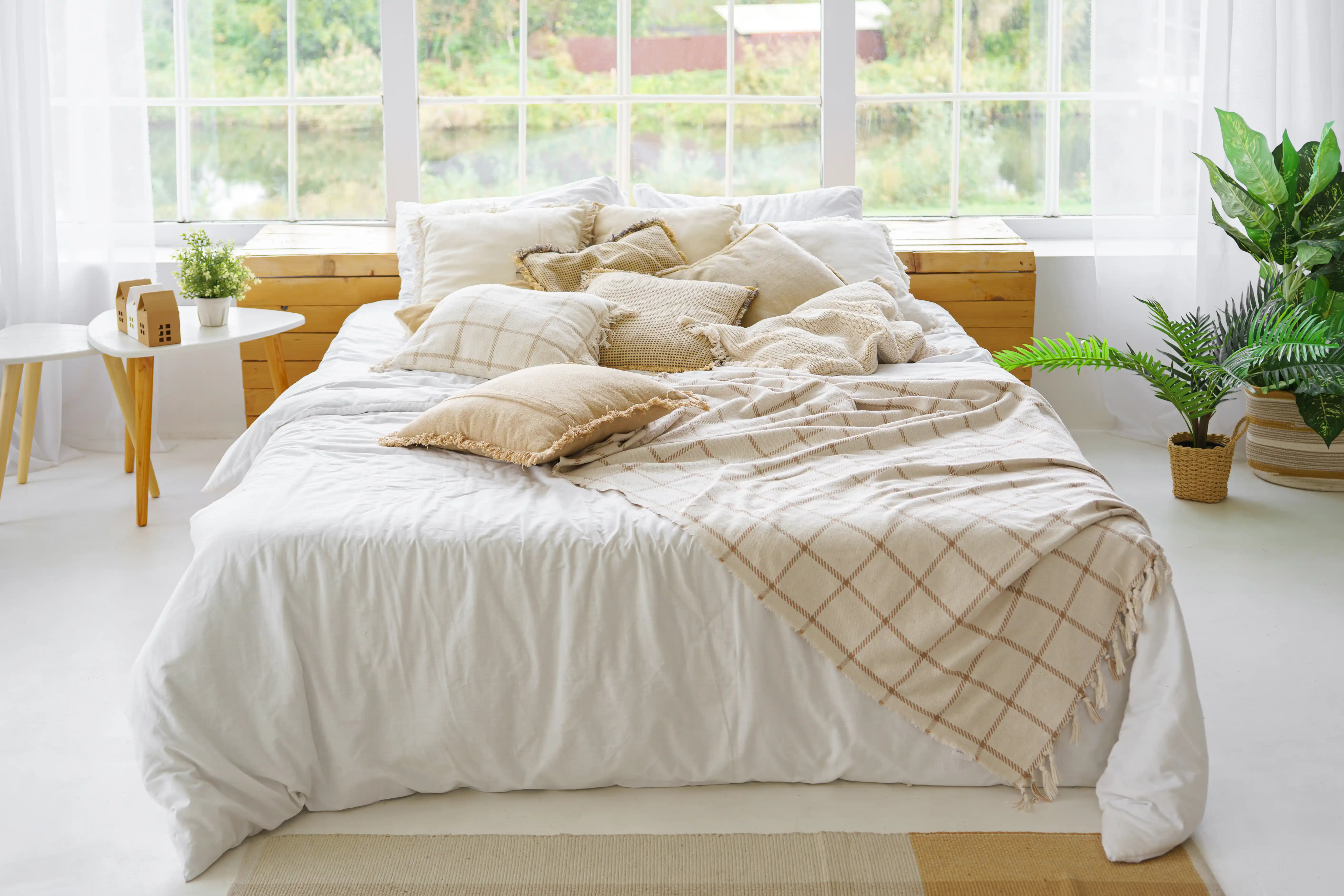 Comfortable bed with lots of soft decorative beige pillows in light sunny bedroom with green plants and large window. Interior design of living room in boho style