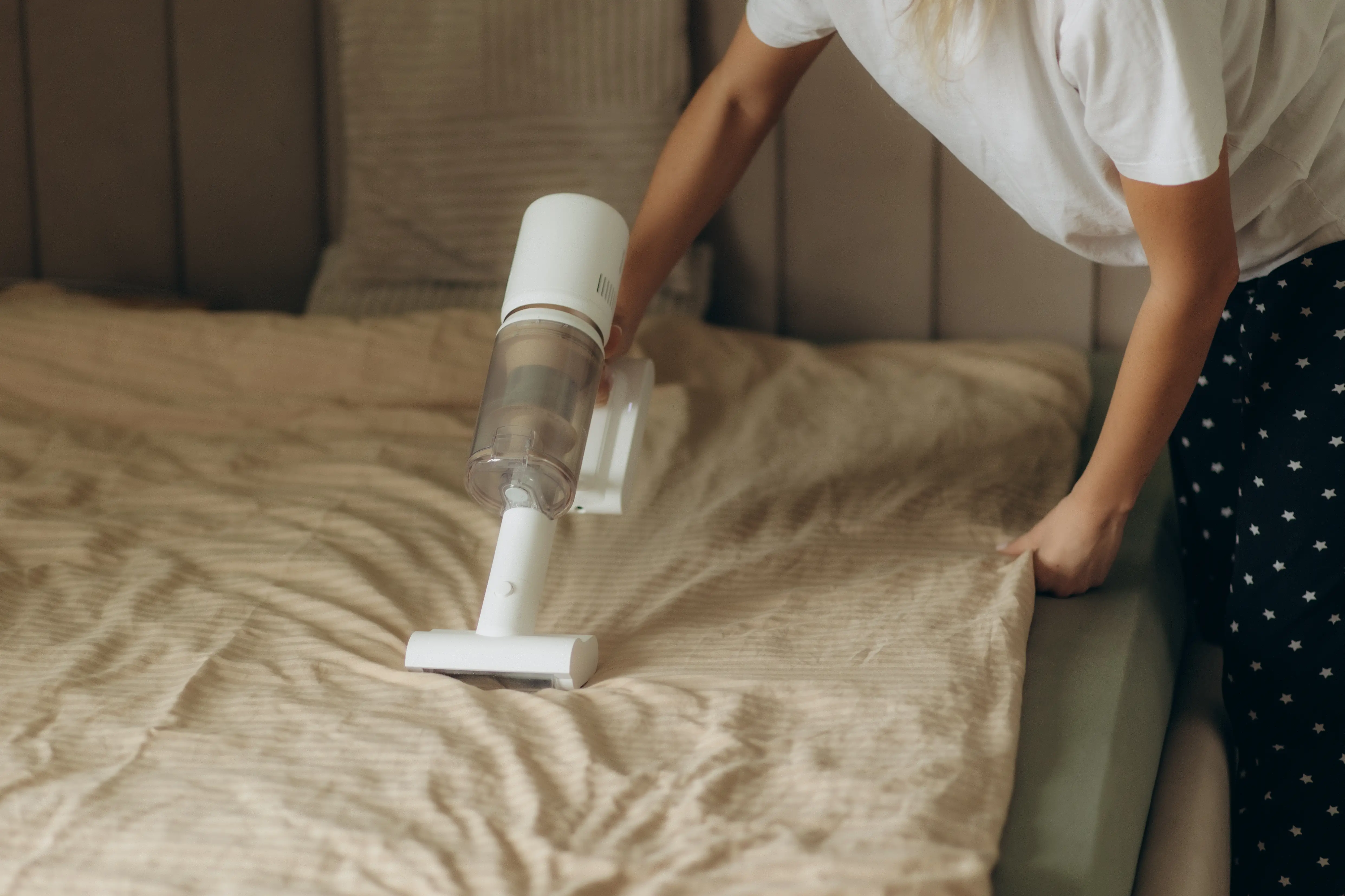 Young asian woman sitting in bed using a wireless vacuum cleaner to clean bedding mites