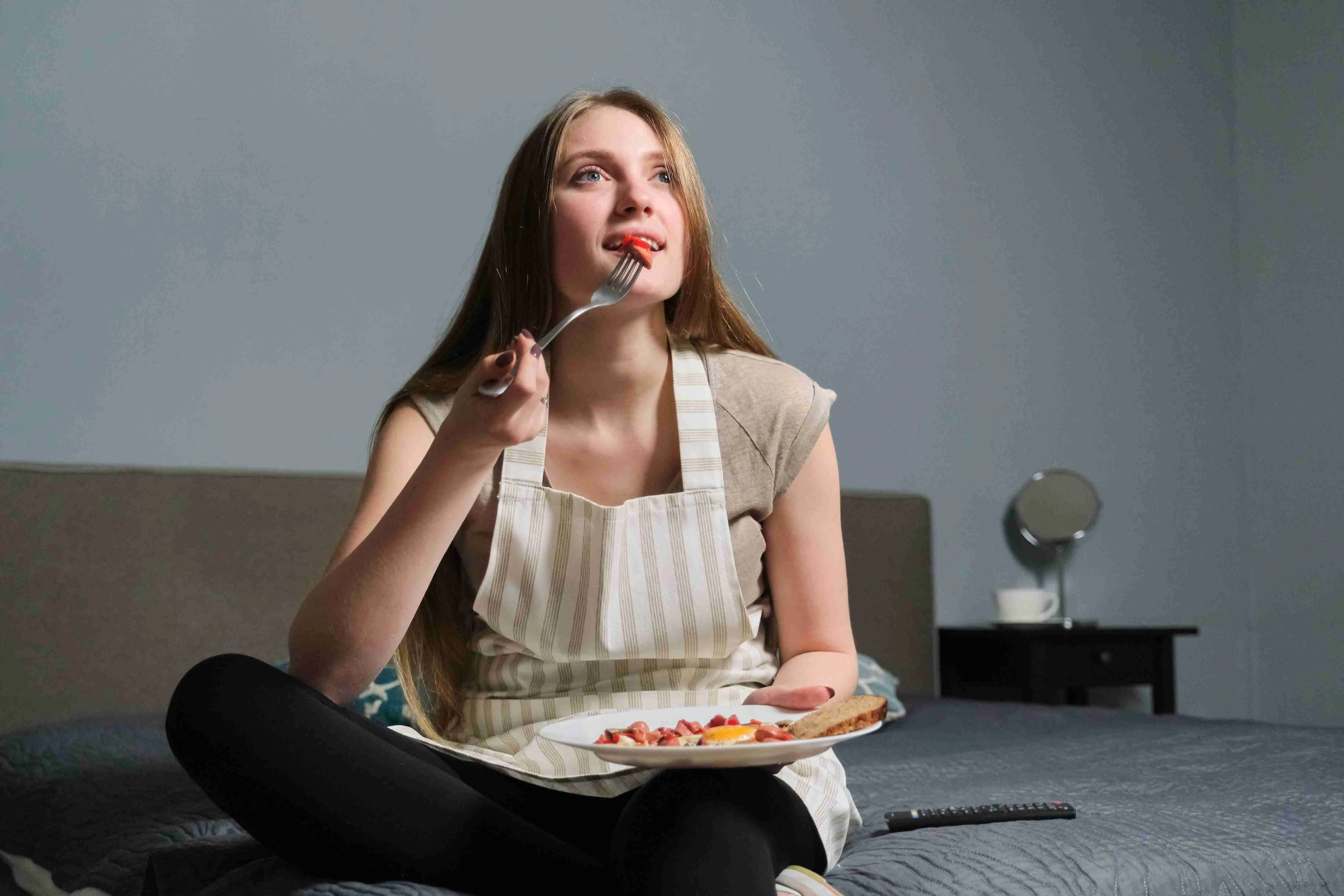 Girl sitting on bed with remote control watching TV and eating