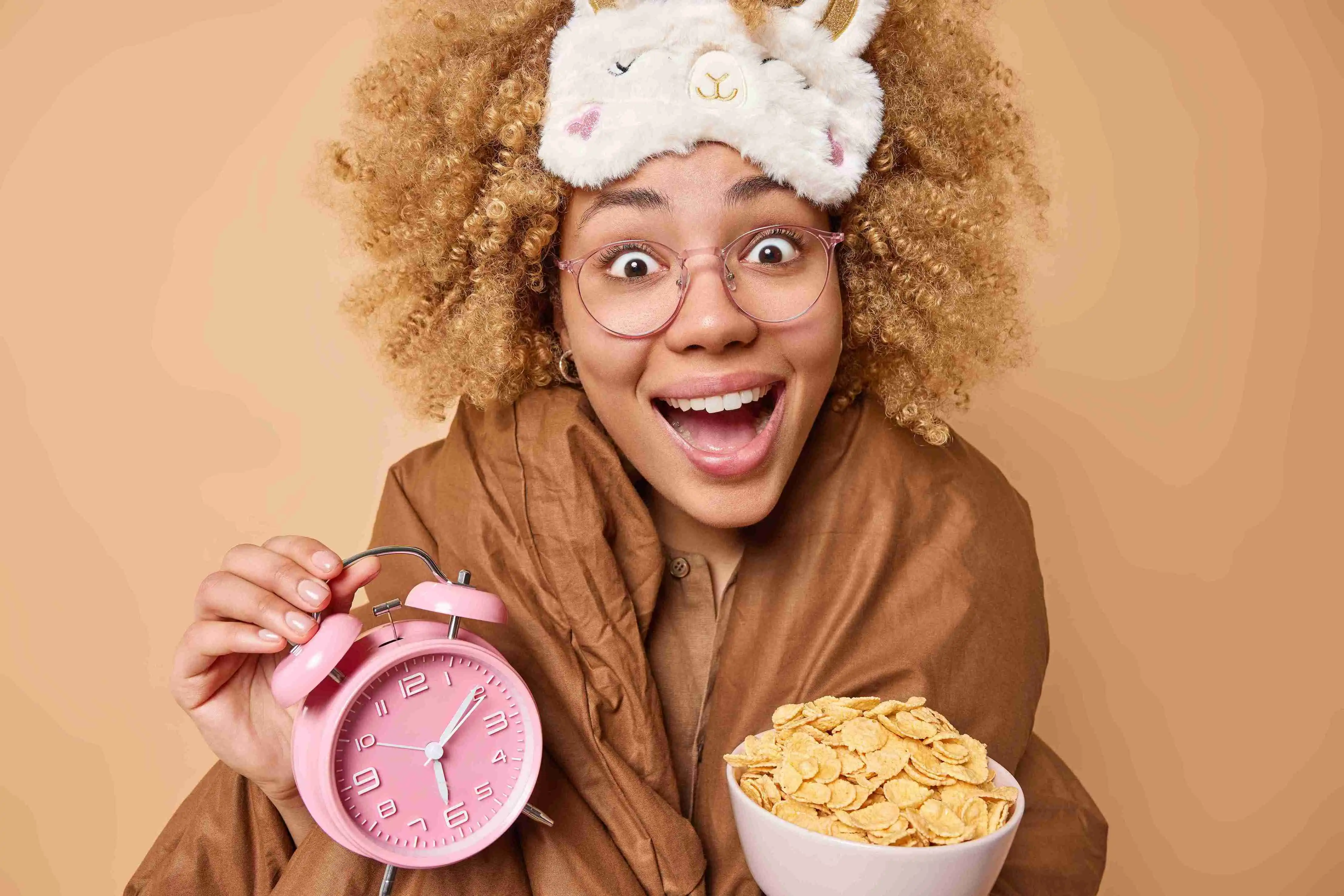 Positive young curly haired woman holds bowl of cereals and alarm clock