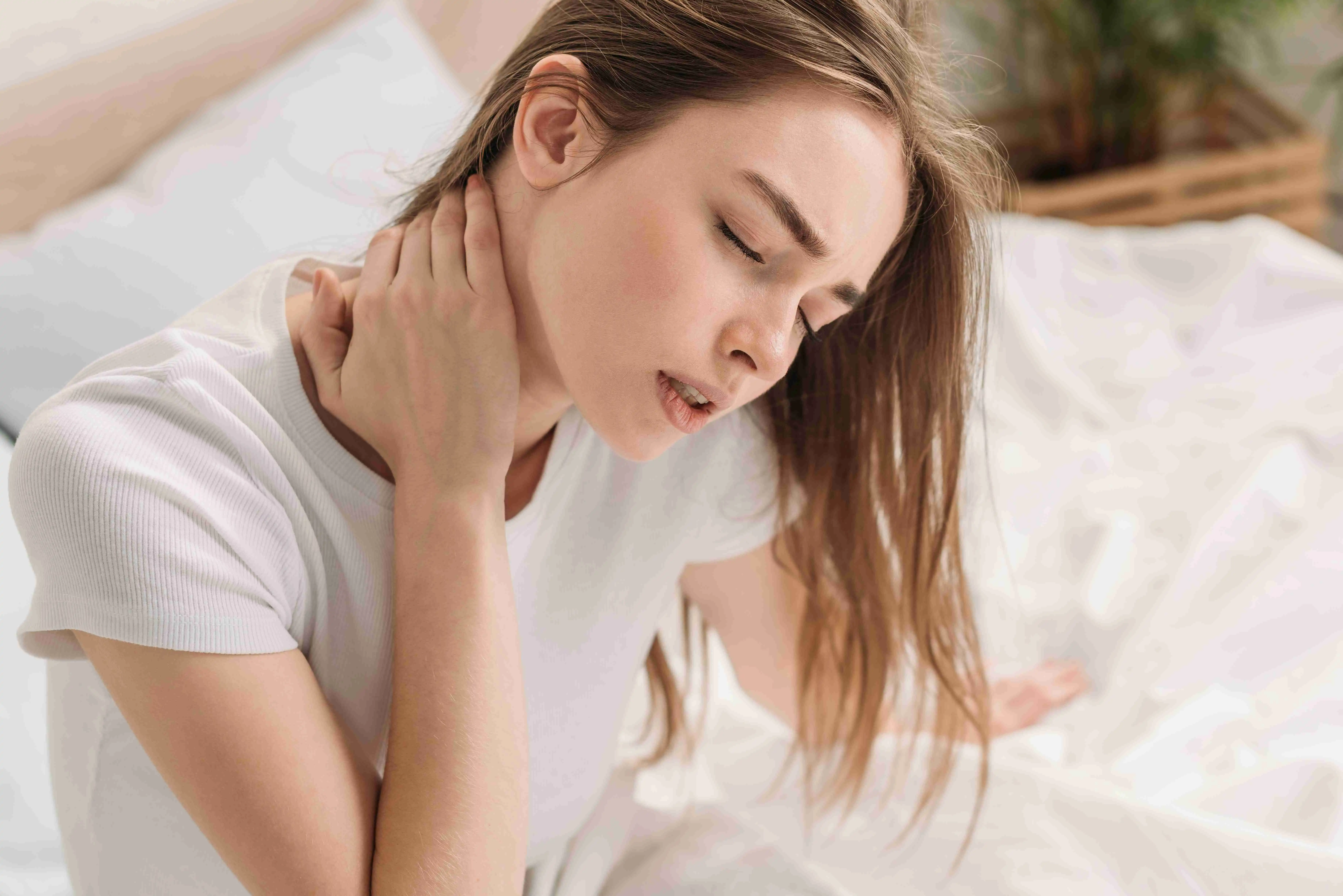 young woman sitting in bed with closed eyes and suffering from neck pain